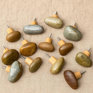 May include: A collection of twelve cabinet knobs, each featuring a smooth, polished river stone in various shades of brown, gray, and green, attached to a light-colored wooden base with a protruding metal screw for installation.