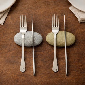 May include: Two forks and two knives are arranged on a wooden surface, each paired with a smooth, gray or green stone. The silverware is stainless steel, and the arrangement suggests a rustic or natural dining theme.