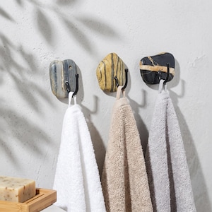 Pode incluir: Três ganchos de parede com bases de pedra natural em cinza, amarelo e preto com veios brancos. Cada gancho tem um braço de metal preto em forma de J. Uma toalha branca, uma bege e uma cinza estão penduradas nos respetivos ganchos.