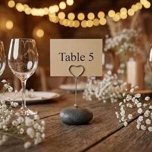 May include: A table number card with the text "Table 5" held by a silver heart-shaped wire. The card sits on a gray stone base. The scene includes wine glasses, white flowers, and soft lighting, suggesting a wedding or event.