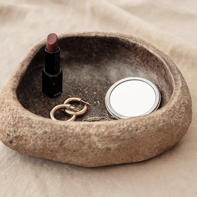 May include: A textured stone bowl contains a lipstick, gold hoop earrings, a small compact mirror, and a delicate gold chain. The lipstick is in a black case with a mauve shade. The earrings are gold and circular. The mirror is round with a silver frame.