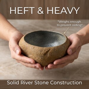 May include: A natural river stone bowl with a textured exterior and a gray interior. The bowl is held in two hands. Text on the image reads "HEFT & HEAVY" and "Solid River Stone Construction". The text also states the bowl weighs enough to prevent sliding.