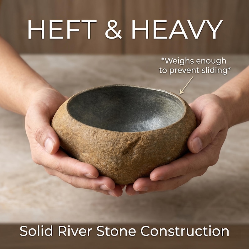 May include: A natural river stone bowl with a textured exterior and a gray interior. The bowl is held in two hands. Text on the image reads "HEFT & HEAVY" and "Solid River Stone Construction". The text also states the bowl weighs enough to prevent sliding.