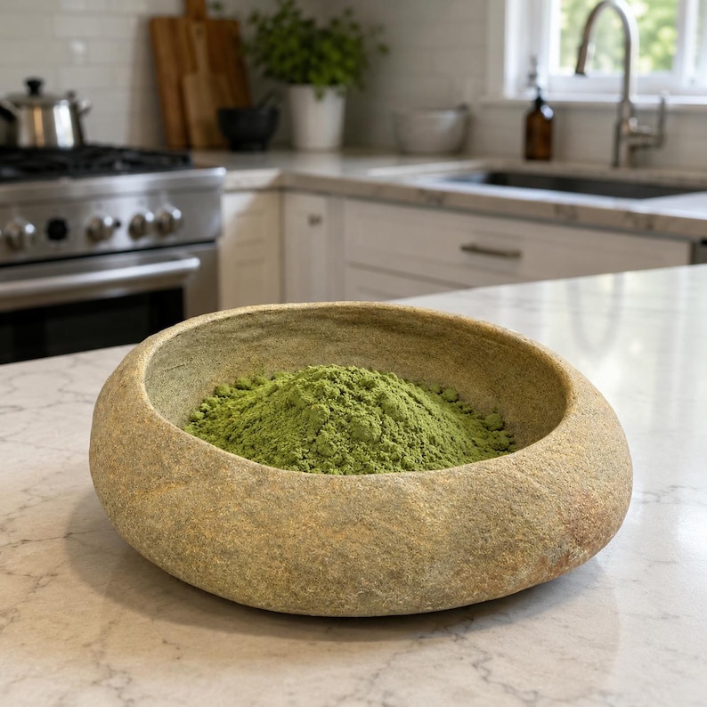 May include: A round, low-profile bowl with a rough, stone-like exterior texture in shades of beige and tan. The interior of the bowl is filled with a mound of fine, vibrant green powder, contrasting with the earthy, natural appearance of the vessel.