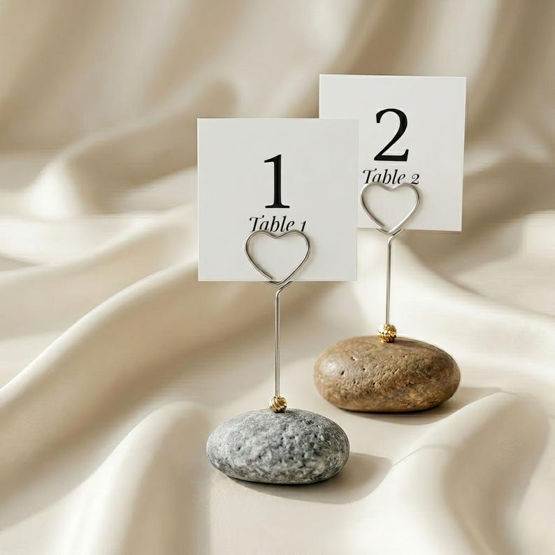 May include: Two place card holders with white cards displaying numbers 1 and 2. The cards are held by silver wire in a heart shape, attached to a small stone base. The stones are gray and brown, with a gold accent.