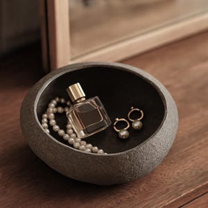 May include: A grey stone-effect bowl contains a perfume bottle, a pearl necklace, and gold earrings with pearl drops. The items are arranged on a wooden surface, suggesting a dressing table. The bowl's dark interior contrasts with the lighter items.