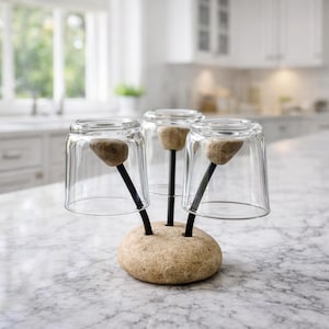 Pode incluir: Um suporte de secagem de vidro exclusivo com três copos transparentes. O suporte apresenta uma base de pedra natural com hastes de metal preto. Cada haste suporta um copo e tem uma pequena pedra arredondada no topo.