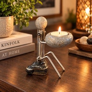 May include: A metal figurine candle holder featuring a smooth, speckled stone head and a larger, rounded stone base that holds a lit tea light candle. The figure has a polished silver-toned metal body with thin, articulated arms and legs.