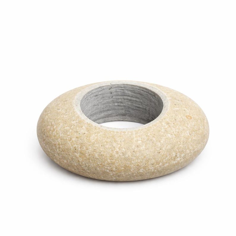 May include: A beige, donut-shaped stone object with a circular opening. The interior of the opening is a light gray color. The stone has a smooth, rounded exterior and a natural, textured appearance.