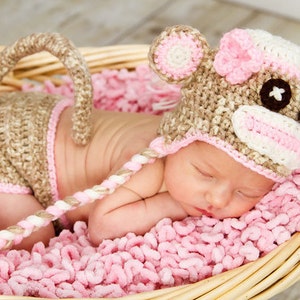 May include: A newborn baby wearing a crocheted monkey hat and diaper cover, lying in a woven basket on a pink blanket. The hat features monkey ears and a face with button eyes. The diaper cover has a tail.
