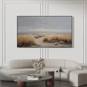 Pode incluir: Uma pintura de paisagem emoldurada retrata uma cena costeira com gramíneas altas e douradas e um caminho arenoso que leva à água. O céu é uma mistura de cinza e azul claro, criando uma atmosfera serena. A obra de arte é exibida acima de um sofá curvo.