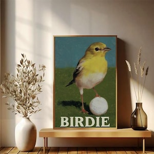 Puede incluir: Impresión artística enmarcada que muestra un pájaro amarillo sobre una pelota de golf. El pájaro se encuentra sobre un fondo verde y azul. La palabra "BIRDIE" está impresa en letras blancas en la parte inferior. La impresión se exhibe en una estantería de madera.