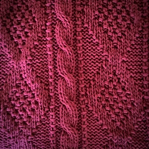 May include: Close-up of a knitted fabric with a cable knit pattern in a deep burgundy color.