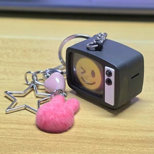 Puede incluir: Un llavero con un televisor en miniatura de color gris oscuro que muestra una cara sonriente amarilla guiñando un ojo en la pantalla. El anillo de metal plateado incluye un dije de estrella, una cuenta de corazón rosa claro y un pompón rosa intenso.