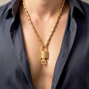 May include: A gold-toned necklace featuring a cylindrical link chain and a rectangular padlock pendant. The padlock displays the LV logo on its front surface and has a small key attached to the bottom. The metal has a polished, reflective finish.