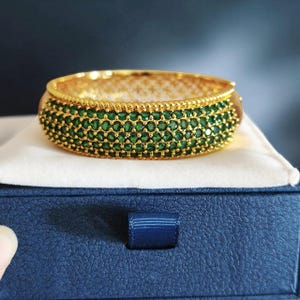 May include: A gold-toned hinged bangle bracelet featuring four horizontal rows of small, round, vibrant green stones set closely together. The edges of the bracelet are finished with a beaded texture, and the interior has a decorative lattice pattern.