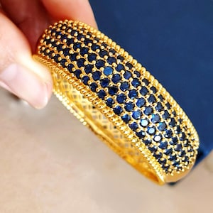 May include: A gold bangle bracelet featuring a wide band encrusted with multiple rows of small, round, deep blue stones. The edges of the bracelet are finished with a textured, beaded gold border, and the inner band displays a lattice pattern.