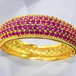 May include: A gold bangle bracelet featuring a wide, curved top surface densely set with multiple rows of small, round, vibrant pinkish-red stones. The edges of the band are detailed with a twisted rope texture, and the inner band has a lattice pattern.