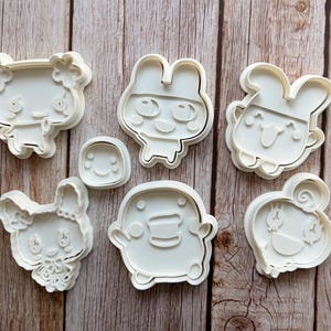以下が含まれることがあります： 様々な漫画風のキャラクターの形をしたオフホワイトのプラスチック製クッキー型6個セット。はっきりとした顔立ちの丸みを帯びた動物のフィギュアや、小さな円形の型が含まれており、木製の表面にグループとして配置されています。