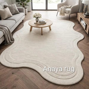 May include: Cream-colored, wavy-edged area rug with a textured surface. The rug features a subtle, layered design along the border. The text "Anaya rug" is visible on the rug. The rug is placed on a wooden floor.