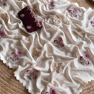 May include: Cream-colored saree with intricate floral embroidery in pink, green, and gold. The fabric has a subtle grid pattern and a scalloped edge. A maroon blouse piece with matching embroidery is included.