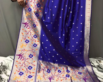 May include: A vibrant blue and silver sari with floral patterns. The sari features a rich blue fabric with small white flower accents. The border is silver with colorful floral and vine designs in blue, yellow, and purple. The sari is draped, showcasing its intricate details.