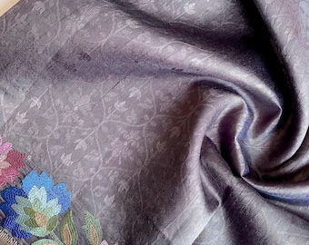 May include: A dark purple fabric with a floral embroidered border. The fabric has a subtle floral pattern throughout. The embroidered border features colorful flowers in shades of pink, blue, and green, adding a decorative touch to the textile.