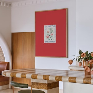 May include: A framed Indonesian postage stamp with a floral design, set against a red background. The stamp is centered within a wooden frame, mounted on a white wall. A green and gold chair sits in front of a striped countertop.