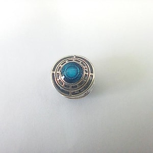 May include: A small, round, silver-toned charm with a detailed design. The center features a vibrant teal-colored enamel, surrounded by a geometric pattern. The charm has a layered appearance, with a textured outer ring.