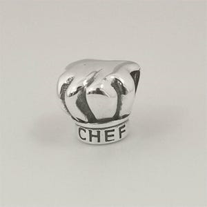 May include: A silver-colored charm in the shape of a chef's hat. The word "CHEF" is engraved on the base of the hat. The charm is detailed with realistic folds and curves, suggesting a three-dimensional design.