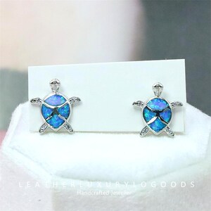 Può includere: Orecchini a lobo a forma di tartaruga color argento con centri blu e verdi simili all'opale e piccole pietre chiare. Gli orecchini sono esposti su una carta bianca.