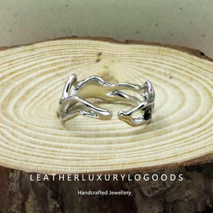 May include: A silver ring with an abstract, flowing design. The ring features an open structure with irregular shapes. It is presented on a wooden surface. The text "LEATHERLUXURYLOGODS" and "Handcrafted Jewellery" are visible.