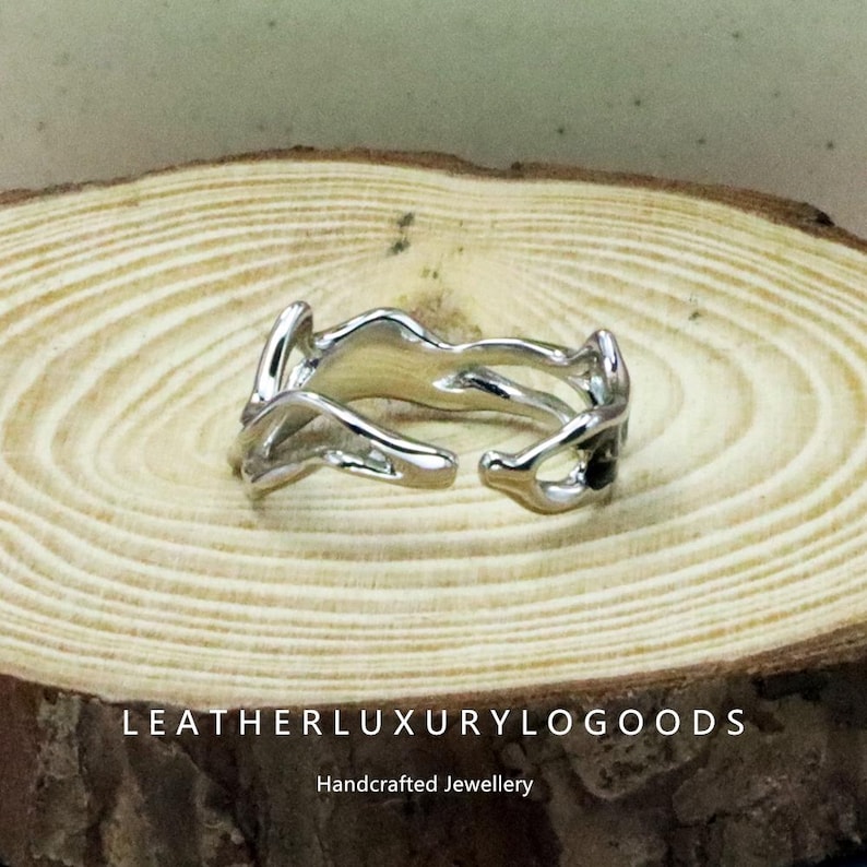 May include: A silver ring with an abstract, flowing design. The ring features an open structure with irregular shapes. It is presented on a wooden surface. The text "LEATHERLUXURYLOGODS" and "Handcrafted Jewellery" are visible.