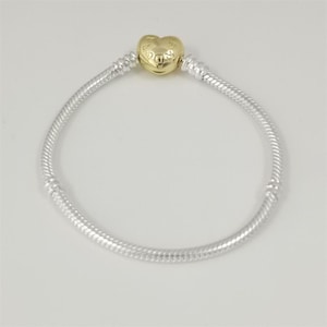 May include: A silver bracelet with a heart-shaped gold charm. The charm has the word "OR" engraved on it. The bracelet has a snake chain design.