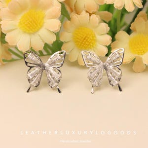 Puede incluir: Dos pendientes de mariposa plateados con detalles de cristal rectangular transparente. Los pendientes se exhiben sobre una superficie beige claro, con flores amarillas y melocotón de fondo. El texto "LEATHERLUXURYLOGOGOODS" está en la parte inferior.
