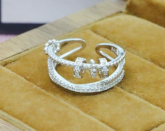 Könnte beinhalten: Ein silberner Ring mit einem einzigartigen Design. Der Ring hat drei Bänder, wobei das mittlere Band drei kleine, rotierende Zylinder aufweist. Das obere und untere Band sind mit kleinen, klaren Steinen besetzt. Der Ring wird auf einer braunen Oberfläche präsentiert.