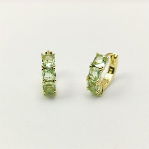 May include: A pair of gold-colored hoop earrings, each adorned with three square, light green gemstones. The earrings are set against a plain white background, highlighting their design and color.