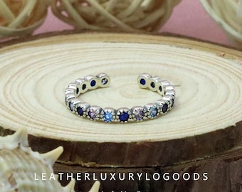 May include: A silver open ring set with small, multi-coloured gemstones, including shades of blue and purple. The ring is displayed on a wooden surface. The text "LEATHERLUXURYLOGGOODS Handcrafted Jewellery" is visible.