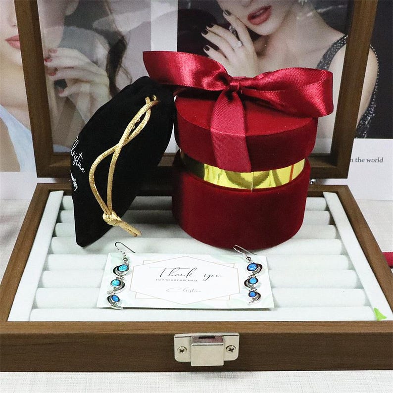 May include: A wooden jewellery display box containing a red velvet gift box with a satin bow, a black velvet pouch, and silver earrings with blue details. A card with "Thank you" is also visible.