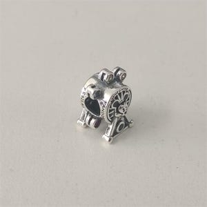 Puede incluir: Un charm plateado con un diseño de noria. El charm tiene detalles intrincados, incluyendo un recorte en forma de corazón y la palabra "PANDORA" grabada en el lateral. La noria tiene un patrón decorativo.