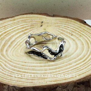 May include: A silver ring with an abstract, flowing design. The ring has an open, irregular shape with a polished finish. The ring is set on a wooden surface. The text "LEATHERLUXURYLOGODS" and "Handcrafted Jewellery" are visible.