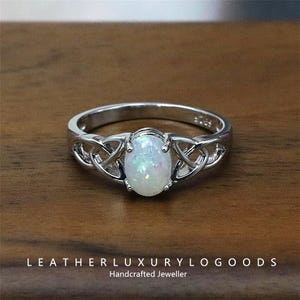 Puede incluir: Un anillo de plata con una piedra central ovalada de ópalo iridiscente. El anillo presenta un diseño de nudo celta en la banda. Las palabras "LEATHERLUXURYLOGGOODS Handcrafted Jeweller" están en la parte inferior de la imagen.