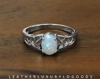 May include: A silver ring with an oval, iridescent opal centre stone. The ring features a Celtic knot design on the band. The words "LEATHERLUXURYLOGGOODS Handcrafted Jeweller" are at the bottom of the image.