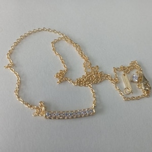 May include: A delicate gold-colored necklace with a bar pendant adorned with small, clear gemstones. The necklace features a fine chain and a small, round gemstone near the clasp. The jewelry is displayed on a light background.