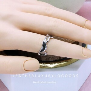 May include: A silver ring with an abstract, flowing design. The ring is worn on a finger, showcasing its unique shape and reflective surface. The ring's design is reminiscent of a wave or ribbon. The text "LEATHERLUXURYLOGODS" and "Handcrafted Jewellery" are visible.