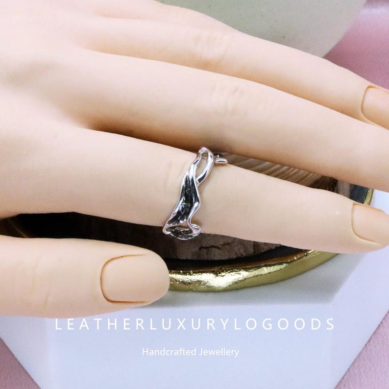 May include: A silver ring with an abstract, flowing design. The ring is worn on a finger, showcasing its unique shape and reflective surface. The ring's design is reminiscent of a wave or ribbon. The text "LEATHERLUXURYLOGODS" and "Handcrafted Jewellery" are visible.