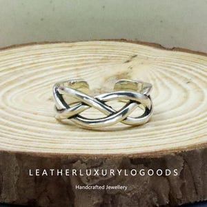 May include: A silver-colored ring with an open design, featuring an interwoven knot pattern. The ring is displayed on a wooden surface. The text "LEATHERLUXURYLOGODS" and "Handcrafted Jewellery" are visible.