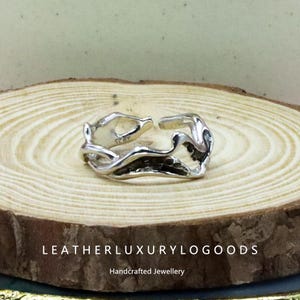 Puede incluir: Un anillo de plata de ley con un diseño orgánico y fluido. El anillo presenta una banda abierta y ajustable y una apariencia texturizada y escultural. El anillo se muestra sobre una superficie de madera. El texto "LEATHERLUXURYLOGGOODS Handcrafted Jewellery" es visible.