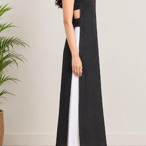 May include: A sleeveless black tunic featuring side cutouts and a long, straight silhouette, paired with wide-leg white trousers. The tunic has a high neckline and extends to the mid-calf, while the white trousers reach the ankles.