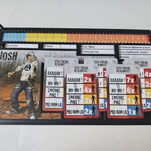 Peut inclure: Un plateau de jeu noir avec une illustration de personnage et des cartes. L'illustration montre une personne avec une arme à feu. Les cartes affichent du texte comme "AAAAAH!" et "HO-HO!". Le plateau a un indicateur de santé.